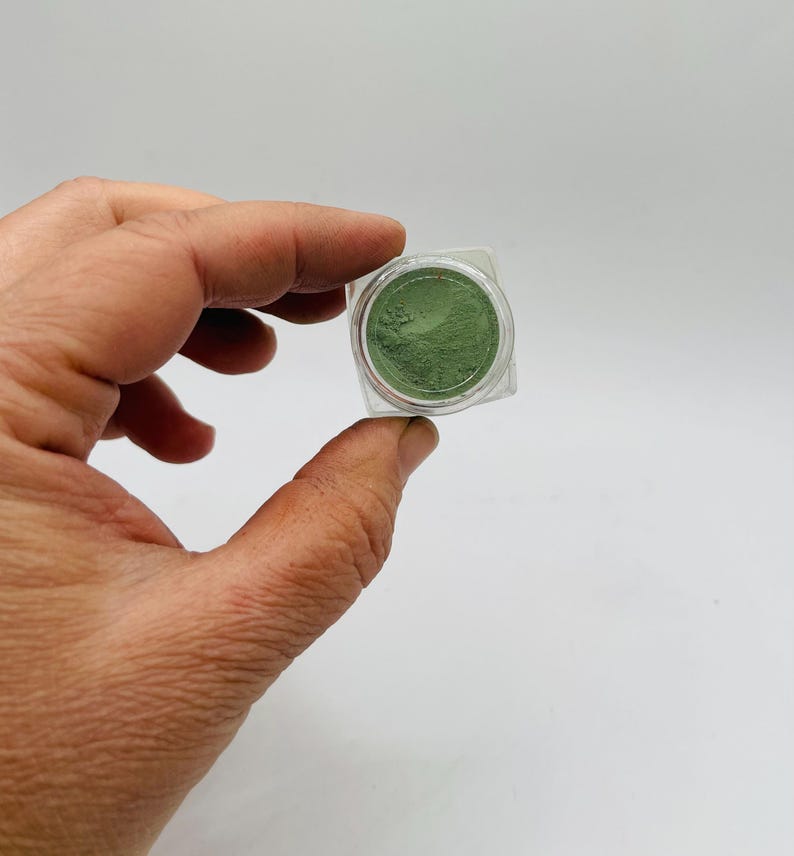 May include: A small, clear plastic container holding a vibrant green powder. The container is square with rounded corners. The powder appears finely milled and evenly distributed within the container. The background is a plain white surface.