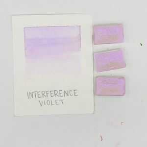 May include: A watercolor swatch of Interference Violet paint, with a gradient from light to dark. Three rectangular paint pans in a similar shade are next to the swatch. The text "INTERFERENCE VIOLET" is written below the swatch.