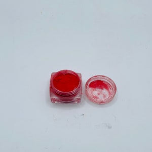 May include: An open, clear, square container filled with bright red powder, next to its clear lid. The container sits on a white surface. The powder appears finely milled, suggesting a cosmetic product like blush or eyeshadow.