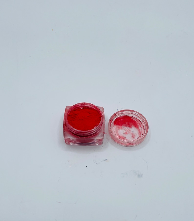 May include: An open, clear, square container filled with bright red powder, next to its clear lid. The container sits on a white surface. The powder appears finely milled, suggesting a cosmetic product like blush or eyeshadow.