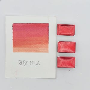 Puede incluir: Una muestra de acuarela con un degradado de tonos rosas y rojos, etiquetada como "RUBY MICA". Tres pastillas de pintura roja metálica rectangulares están colocadas a la derecha. La imagen está sobre un fondo blanco.