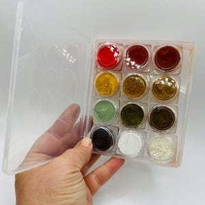 May include: A clear plastic case holding twelve small jars of colorful powders. The jars are arranged in three rows of four, with colors including red, yellow, green, and brown. The case is being held by a hand.