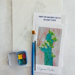 Op de afbeelding: Een verf-op-nummer-set met een Saguaro-cactus. De set bevat een blanco vel papier met een potloodschets van de cactus, een referentiebeeld van een geschilderde cactus, een set van vier waterverfverven, twee kwasten en instructies over hoe je de cactus in zes eenvoudige stappen kunt schilderen.