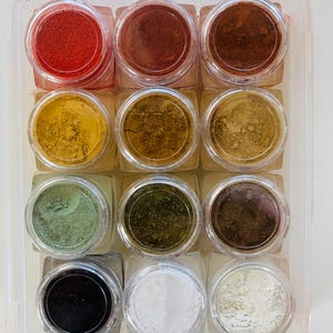 May include: A clear plastic container holding twelve small jars of colorful powders. The powders range in color from red and yellow to green and black. The jars are arranged in three rows of four.