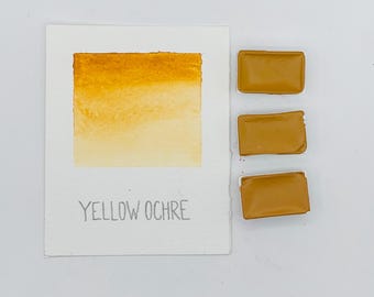 May include: A watercolor swatch of yellow ochre paint with a gradient effect, next to three rectangular paint pans. The text "YELLOW OCHRE" is printed below the swatch. The paint colors are shades of yellow and brown.