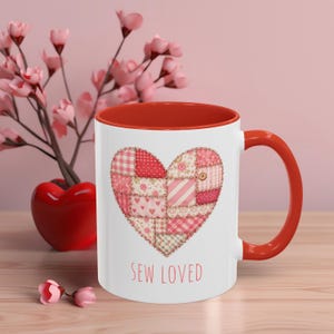 May include: White ceramic mug with a red interior and handle. The mug features a patchwork heart design in shades of pink and red, with the words "SEW LOVED" in red script below the heart. A decorative item for sewing enthusiasts.