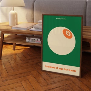 May include: A green and white poster with a brown frame. The poster features a white billiard ball with an orange number eight on it. The text "Sunday Studios" and "Leave it up to luck" are printed on the poster.