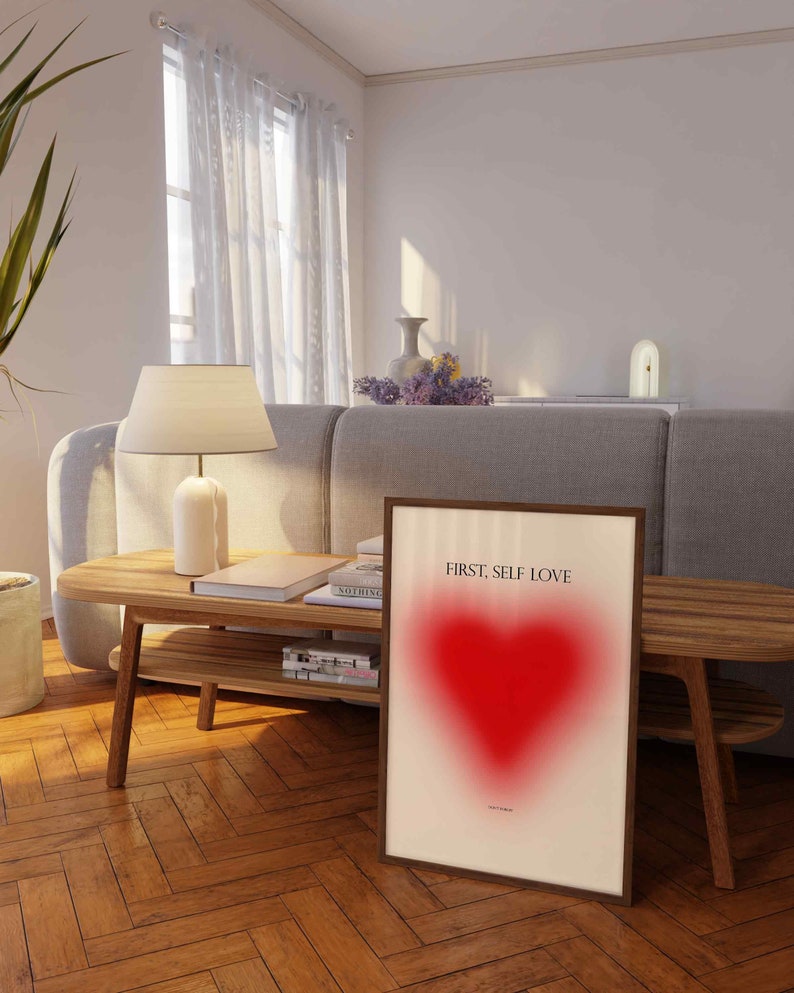 May include: A framed print with a red heart and the text "FIRST, SELF LOVE" on a white background. The print is leaning against a wooden table in a living room setting.