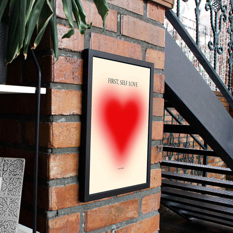 May include: A black framed poster with a white background and a red heart in the center. The text "FIRST, SELF LOVE" is printed in black at the top of the poster.