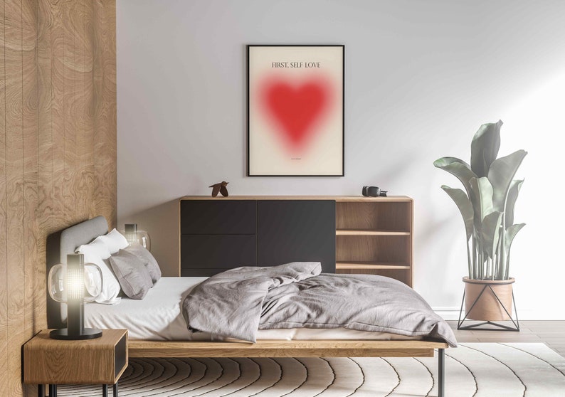 May include: A bedroom interior with a bed, a wooden nightstand, a lamp, a dresser, a plant, and a framed print with a red heart and the text "FIRST, SELF LOVE".