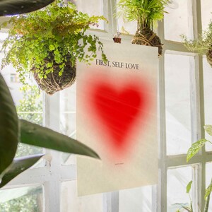 May include: A white poster with the text "FIRST, SELF LOVE" printed in black. A red heart shape is in the center of the poster.