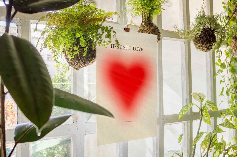 May include: A white poster with the text "FIRST, SELF LOVE" printed in black. A red heart shape is in the center of the poster.