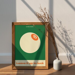 May include: A framed poster with a green background and a white 8-ball with an orange number 8. The text "sunday studios" and "Leave it up to luck" are printed in white on the poster.