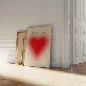 May include: A white poster with a gold frame featuring a red heart with a blurred effect. The poster has the text "FIRST, SELF LOVE" at the top.
