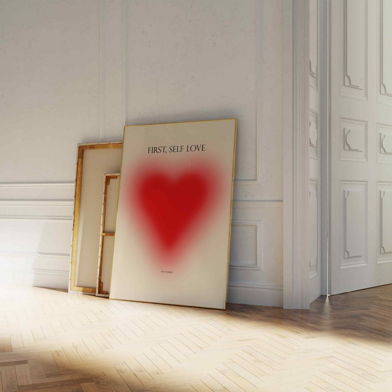 May include: A white poster with a gold frame featuring a red heart with a blurred effect. The poster has the text "FIRST, SELF LOVE" at the top.