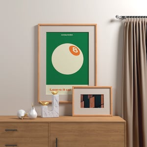 May include: A framed poster with a green background and a white 8-ball with an orange number 8. The poster has the text "sunday studios" and "Leave it up" in white. A smaller framed picture with a brown and black abstract design is below the poster. The posters are on a wooden dresser with two white ceramic pears and a gold metal bird sculpture.