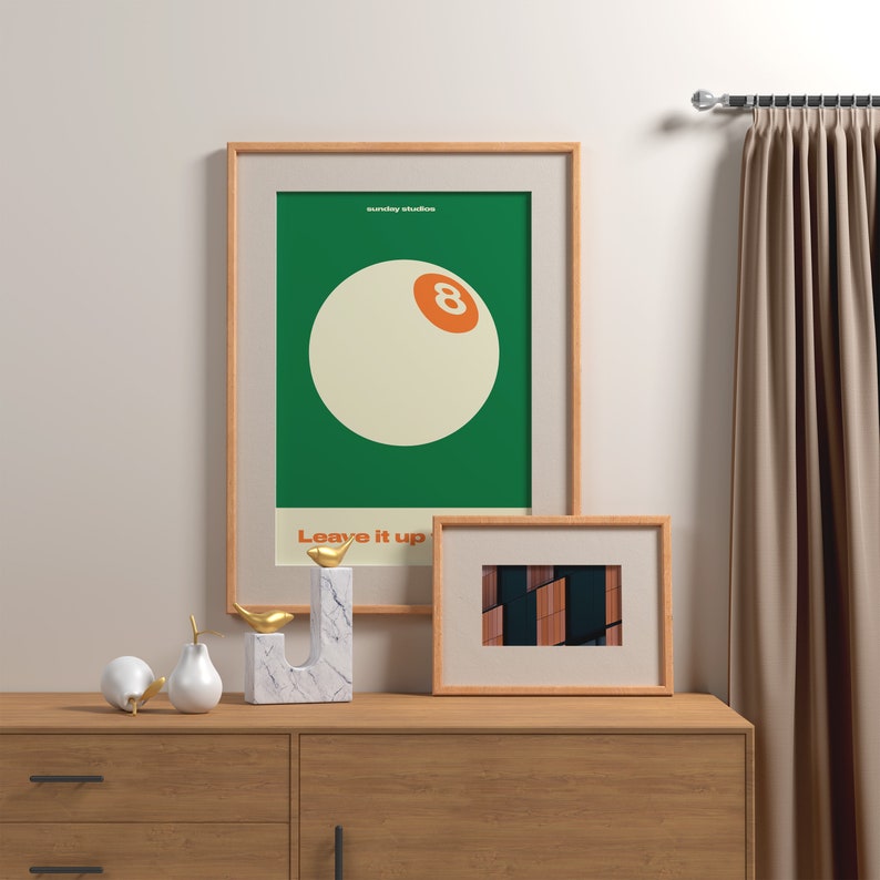 May include: A framed poster with a green background and a white 8-ball with an orange number 8. The poster has the text "sunday studios" and "Leave it up" in white. A smaller framed picture with a brown and black abstract design is below the poster. The posters are on a wooden dresser with two white ceramic pears and a gold metal bird sculpture.