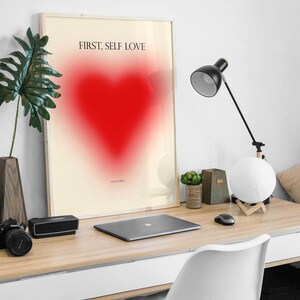 May include: A framed print with a white background and a red heart in the center. The text "FIRST, SELF LOVE" is printed in black at the top of the print.
