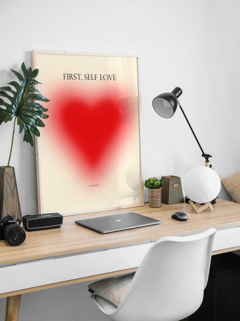 May include: A framed print with a white background and a red heart in the center. The text "FIRST, SELF LOVE" is printed in black at the top of the print.