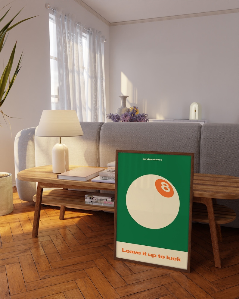 May include: A green and white poster with a brown frame. The poster features a white billiard ball with an orange number eight on it. The text "Sunday Studios" and "Leave it up to luck" are printed on the poster.