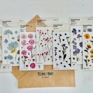 May include: A collection of pressed flower stickers in various floral designs, including daisies, hydrangeas, and cosmos. The stickers are displayed in clear packaging, with a brown envelope in the foreground. The text "Pressed Flower Sticker" is visible.