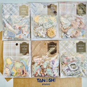 May include: Six packs of decorative stickers in various designs, including flowers, animals, and architectural elements. Each pack is sealed in clear plastic and labeled "Little & Dear". The bottom right corner of the image has the text "TANOSHI STUDIO".
