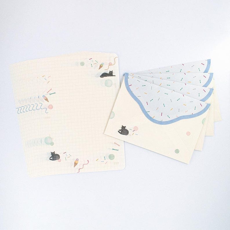 May include: A set of stationery featuring a black cat and ice cream cone design. The paper is white with a grid pattern and the envelopes have a blue scalloped edge with a confetti pattern.