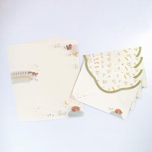 May include: A set of stationery featuring a cream-colored envelope with a green scalloped edge and a cream-colored sheet of paper with a grid pattern. The envelope and paper are decorated with a floral pattern and illustrations of squirrels and a sleeping fox.