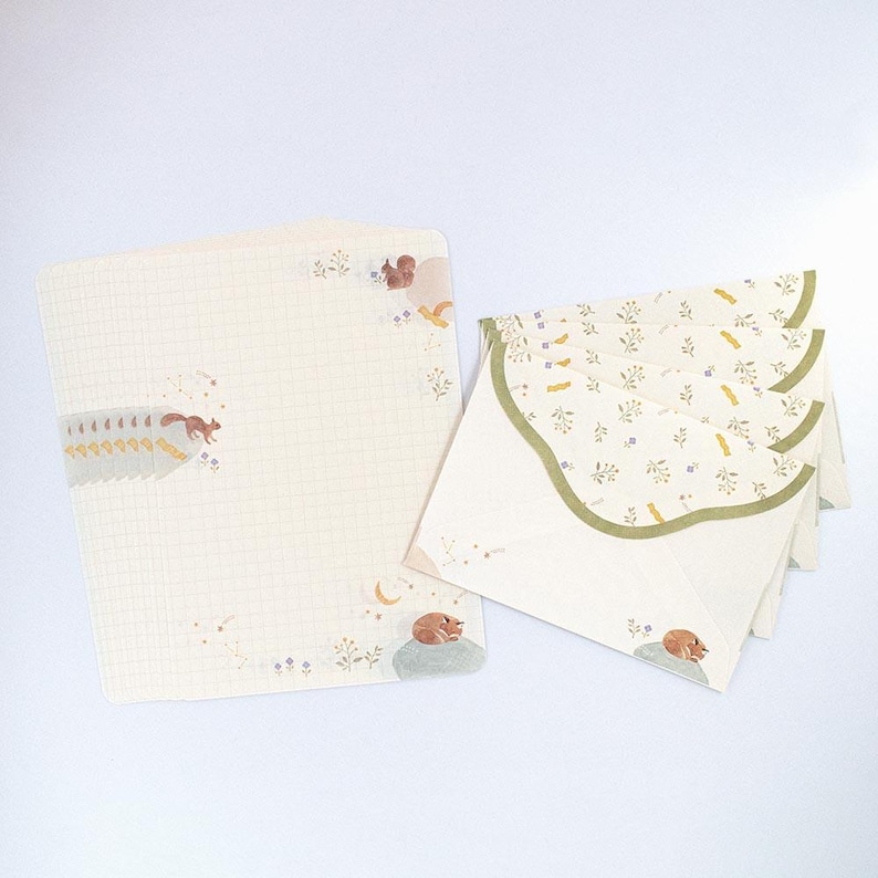 May include: A set of stationery featuring a cream-colored envelope with a green scalloped edge and a cream-colored sheet of paper with a grid pattern. The envelope and paper are decorated with a floral pattern and illustrations of squirrels and a sleeping fox.