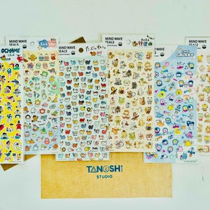 May include: A collection of Mind Wave Seals sticker sheets featuring various designs. The sheets showcase colorful illustrations of people, animals, and space-themed characters. The Tanoshi Studio logo is visible on a brown card.