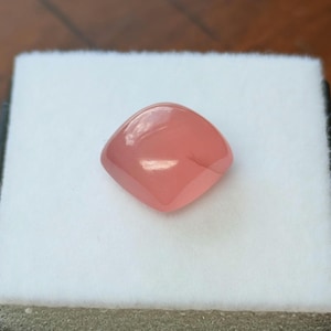 May include: A square, polished gemstone with rounded corners. The gem is a soft, translucent pink color, set against a white background. The gemstone's surface reflects light, giving it a glossy appearance. The gem is likely a cabochon, suitable for jewelry.