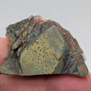 Może przedstawiać: Zbliżenie okazu mineralnego o warstwowej strukturze. Okaz charakteryzuje się mieszanką kolorów, w tym zieleni, złota, brązu i bieli. Powierzchnia ma teksturowany, krystaliczny wygląd z małymi, błyszczącymi wtrąceniami.