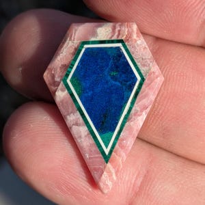 May include: A pentagonal gemstone inlay featuring a central deep blue azurite stone surrounded by a thin white border and a green malachite frame, all set within a textured, light pink rhodochrosite stone with a polished, flat surface.