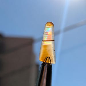 May include: A small, iridescent, brown and orange agate gemstone with a pointed tip. The gemstone is held in a metal clamp.