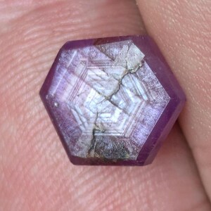 May include: A faceted, hexagonal gemstone with a deep magenta hue. The gem has internal geometric patterns and a visible fracture across its surface. The stone is held between fingers, showcasing its size and color.