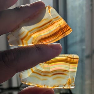 Puede incluir: Dos piezas de ágata con un patrón en capas y bandas en tonos de blanco, naranja y marrón. La ágata es translúcida y muestra colores iridiscentes a la luz.