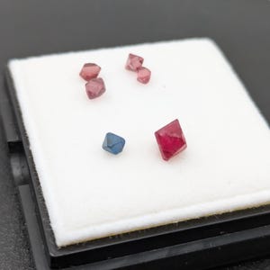 May include: A collection of small, faceted gemstones in shades of red and blue, displayed on a white surface. The gemstones have a geometric shape and are arranged in a scattered pattern. The gemstones are likely raw or unpolished. The background is black.
