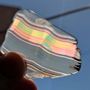 May include: A translucent, iridescent agate slice with a layered pattern of pink, yellow, green, and black bands. The agate is held in a hand against a blue sky background.
