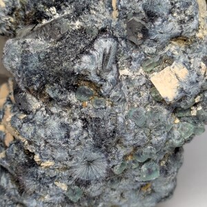 May include: Close-up of a mineral specimen with a complex, textured surface. The rock features a mix of dark gray, teal, and off-white colors. Small, crystalline structures and clusters are visible throughout the specimen, creating a detailed and intricate appearance.
