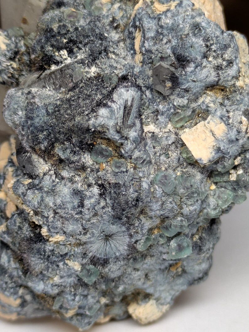 May include: Close-up of a mineral specimen with a complex, textured surface. The rock features a mix of dark gray, teal, and off-white colors. Small, crystalline structures and clusters are visible throughout the specimen, creating a detailed and intricate appearance.