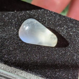May include: A white, teardrop-shaped moonstone cabochon with a smooth, polished surface. The stone is set against a black background.