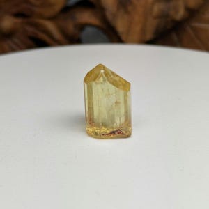 May include: A yellow crystal with a pointed top and a clear, translucent appearance. The crystal has a slightly rough texture and is set against a white background.