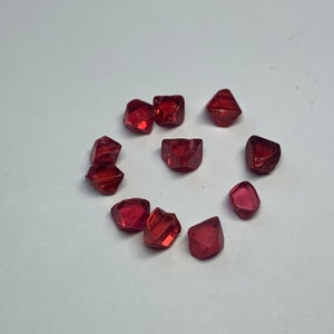 May include: A collection of ten red, faceted, gemstone crystals arranged in a circle on a white surface.