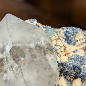 May include: Close-up of a clear quartz crystal with a pointed tip, next to a cluster of small, light-colored crystals and dark blue-green mineral formations. The quartz has a slightly cloudy appearance with some orange and brown inclusions.