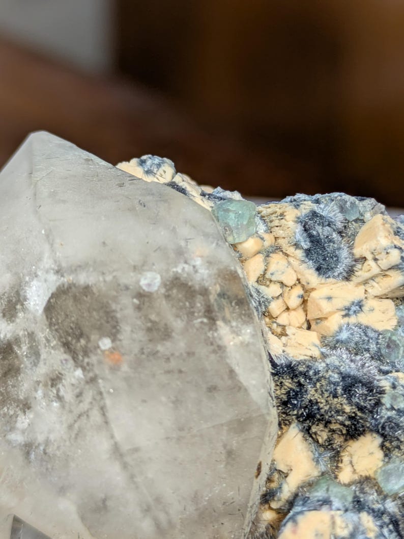 May include: Close-up of a clear quartz crystal with a pointed tip, next to a cluster of small, light-colored crystals and dark blue-green mineral formations. The quartz has a slightly cloudy appearance with some orange and brown inclusions.