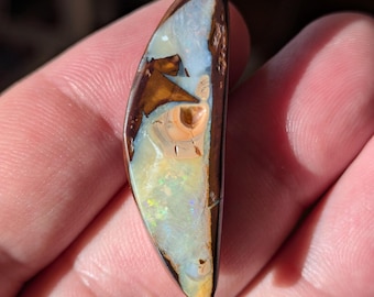May include: A polished opal gemstone with a brown, tan, and blue color scheme. The opal has a unique pattern with iridescent flashes of color. The edges are a dark brown, and the stone is held in a hand.