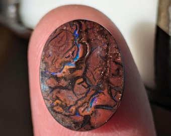 May include: Oval-shaped opal gemstone with a dark brown base color and intricate patterns. The opal displays flashes of blue, orange, and yellow, creating a unique and colorful appearance. The gemstone is held by a finger.