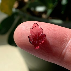 May include: A small, carved, red leaf made of glass. The leaf is translucent and has a detailed design.