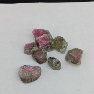 Peut inclure: Un ensemble de cristaux de tourmaline bruts aux tons roses, verts et gris. Les pierres naturelles et non polies présentent des tailles et des formes variées, avec des textures et des dégradés de couleurs visibles. Les cristaux sont disposés sur une surface blanche.