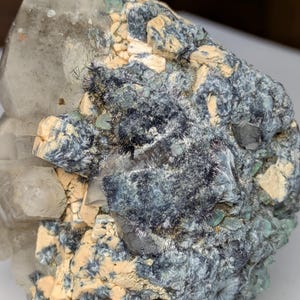 May include: A close-up of a natural mineral specimen. The crystal formation features a large, clear quartz crystal with a dark, textured matrix. The matrix has areas of blue-grey, tan, and green hues, creating a striking contrast.